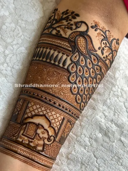 A close-up of a bridal arm design, featuring a large peacock surrounded by trees and a band of elephants.