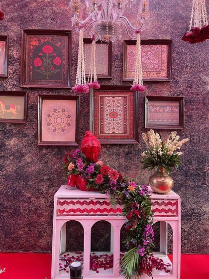 A detail shot from the red and gold Shreenathji wedding, showing a beautifully decorated corner. A console with traditional arches is adorned with a floral runner and framed artwork, showcasing how every element is curated to fit the grand theme.