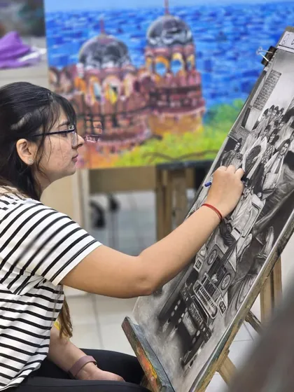A student is captured in a moment of deep concentration, working on her complex charcoal scene. The background shows other artworks, highlighting the inspiring environment of our studio.