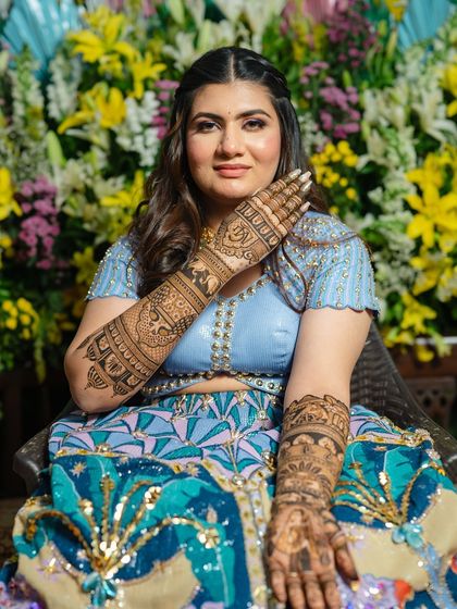 A stunning bride posing against a floral backdrop. Her full-sleeve mehendi is a work of art that perfectly matches her vibrant blue lehenga.