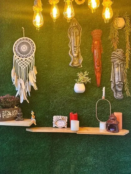 A night-time shot of the green wall balcony. The warm Edison bulbs hanging from the ceiling highlight the textures of the dreamcatcher, ethnic masks, and plants, creating a super cosy and inviting atmosphere.