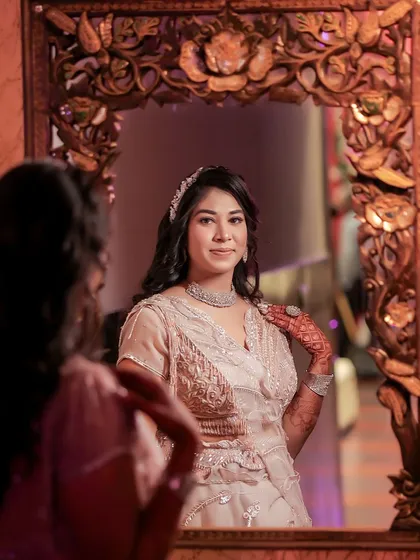 A creative portrait of the bride looking at her reflection in an ornate mirror. This shot captures her from two angles, highlighting her elegant hairstyle, jewelry, and happy expression.