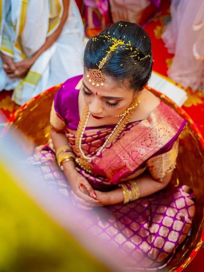 An overhead shot of a South Indian bride during a wedding ritual, highlighting her stunning purple silk saree and traditional gold jewelry.