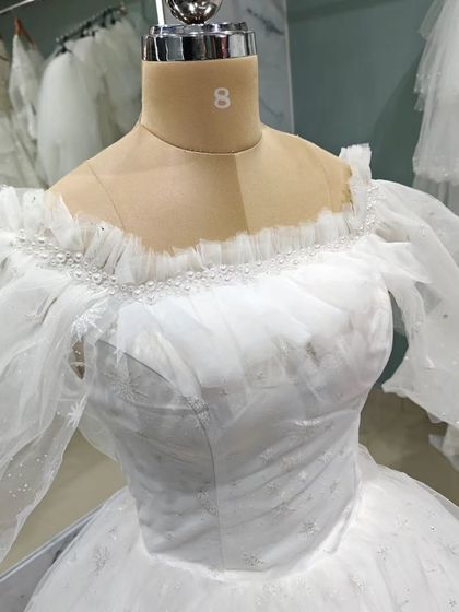 The bodice of this fairytale gown features a delicate off-shoulder neckline with ruffled tulle and pearl details. It's a romantic design for a bride who wants to feel like she's stepped out of a storybook.