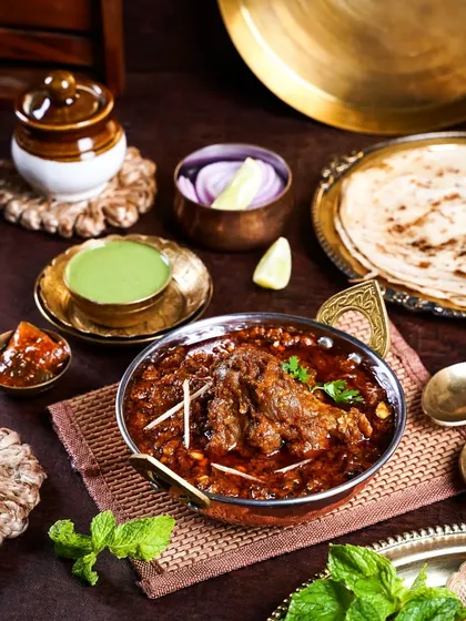 A rich and hearty mutton curry in a traditional copper bowl. The dark, moody setup with rustic props creates a warm and inviting atmosphere, perfect for showcasing robust Indian flavors.