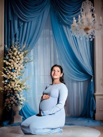 A serene portrait in a light blue gown. The mom-to-be is kneeling against a grand backdrop with blue curtains and a crystal chandelier, creating a regal and elegant atmosphere.