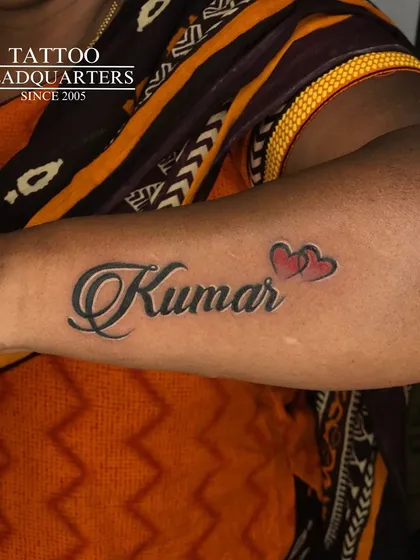 The name "Kumar" in a bold, shaded script with two small red hearts, creating a classic and loving name tattoo.