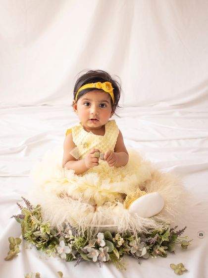 A sweet baby girl in a frilly yellow dress sits in a bowl decorated with flowers. A perfect portrait for a six-month milestone.