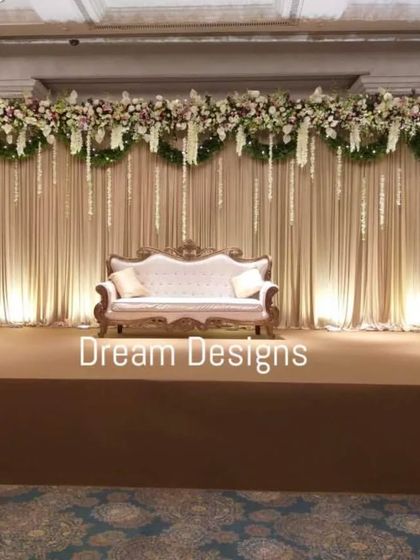 A classic and elegant reception stage design. The backdrop of soft, draped fabric is accented with a lush floral runner and hanging white blossoms, creating a timeless and sophisticated setting.