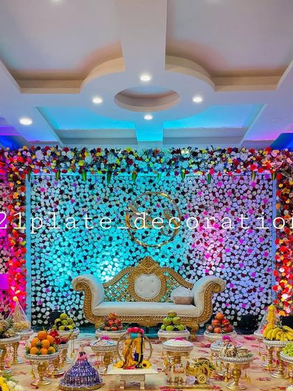 A grand baby shower decoration with a full floral wall and dramatic lighting. The setup includes a beautiful sofa and a complete display of traditional plates and dolls.