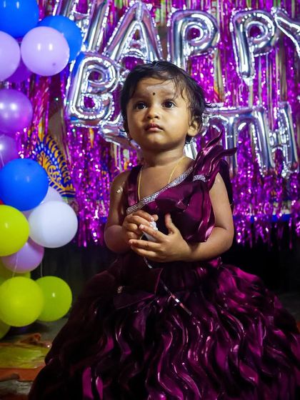 The birthday girl looking regal in her beautiful purple dress. The shiny "Happy Birthday" balloons and colorful party decor create a vibrant and celebratory setting for her first birthday portrait.