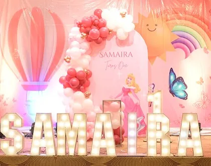 A charming princess theme for Samaira's first birthday, featuring a pink backdrop with a hot air balloon, a smiling sun, and Princess Aurora. The marquee letters add a touch of glamour.