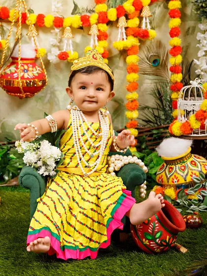 A playful little Radha kicks up her foot while sitting on a miniature green chair in the festive Janmashtami setting.