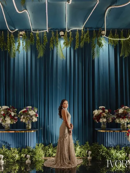 A bride stands poised against a dramatic blue and green backdrop, her elegant gown contrasting beautifully with the modern, stylish decor of the Sangeet venue.