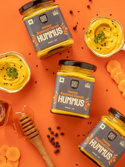A top down shot of Honey Roasted Carrot Hummus. The styling includes the key ingredients like carrots and honey, which visually tells the story of the product's flavor. The color palette is warm and inviting.