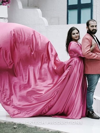 A couple poses against a white, Santorini-style building, the bride's pink trail gown billowing dramatically for a high-fashion pre-wedding look.