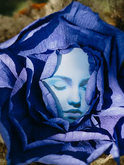 A surreal art piece featuring a serene face nestled within the petals of a deep blue paper flower, blending art, nature, and a touch of the fantastical.