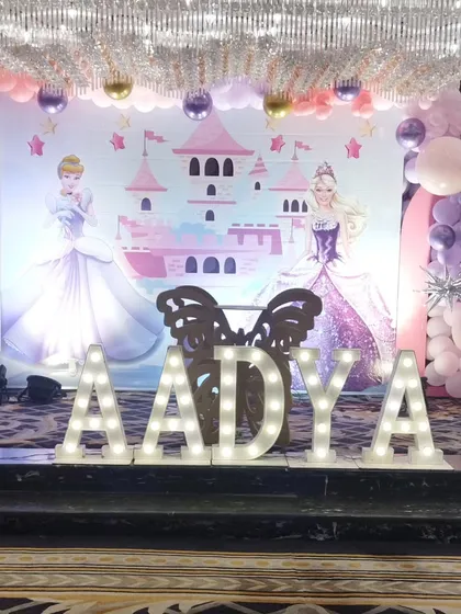 For a magical princess party, I designed this beautiful stage featuring a castle backdrop and two Disney princesses. The large, illuminated marquee letters for the name 'AADYA' create a perfect centerpiece for photos, complemented by soft pink and purple balloon decorations.