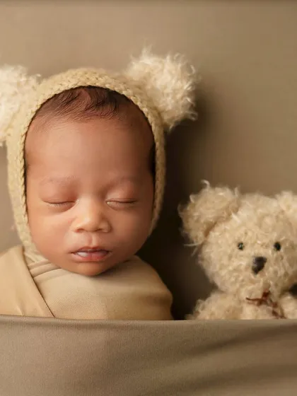 A close-up portrait of a newborn in a bear bonnet, sleeping soundly next to a teddy bear. The neutral, earthy tones create a warm and timeless feel for these infant photos.