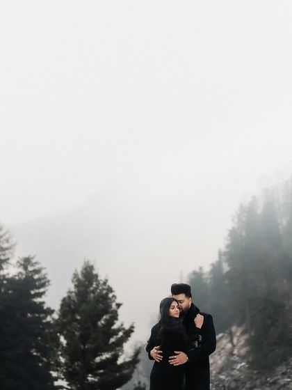 A couple framed by pine trees against a foggy mountain backdrop. The moody atmosphere and intimate pose create a deeply romantic and cinematic pre-wedding photograph.