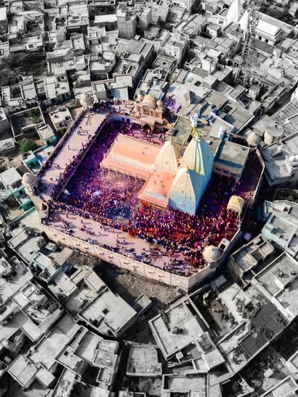 A color-splash version of the aerial Holi photo, with the temple and celebration in full color while the surrounding city is in black and white, emphasizing the festive energy.