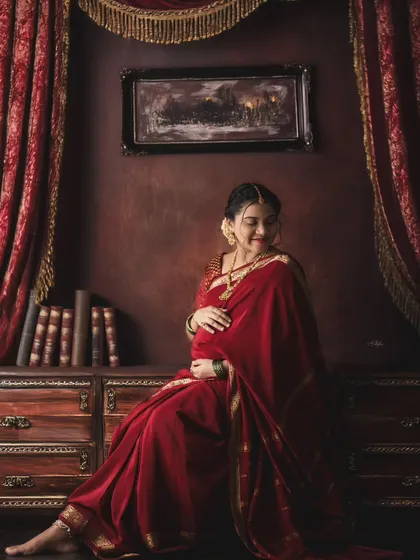 A regal and timeless portrait of a mother-to-be in a rich red saree. The vintage-inspired studio setup with dark wood and classic decor adds a touch of royal elegance.