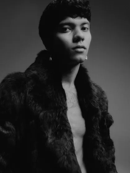 A black and white editorial portrait featuring a male model in a fur coat. The lighting is dramatic and moody, creating a sense of luxury and mystery.