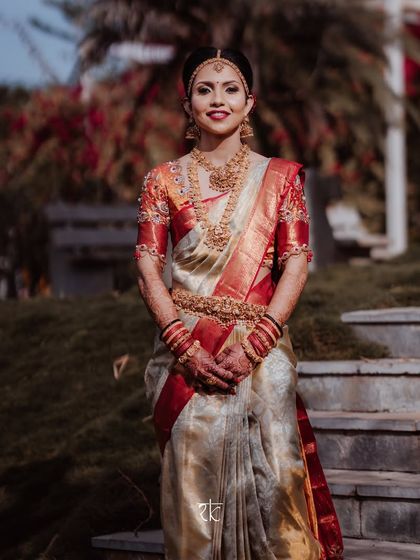 Our bride stands poised and elegant in a classic off-white and red Kanjeevaram saree. The custom blouse, with its intricate sleeve embroidery, perfectly complements this timeless Muhurtham look.