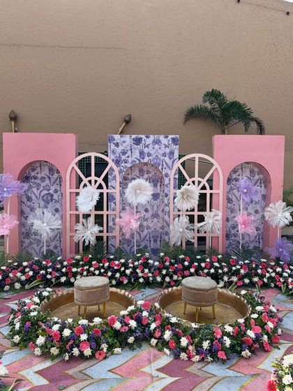 A whimsical pastel Haldi stage we built in the middle of a pool. The backdrop features pink arched panels, floral-patterned wallpaper, and delicate paper flowers.