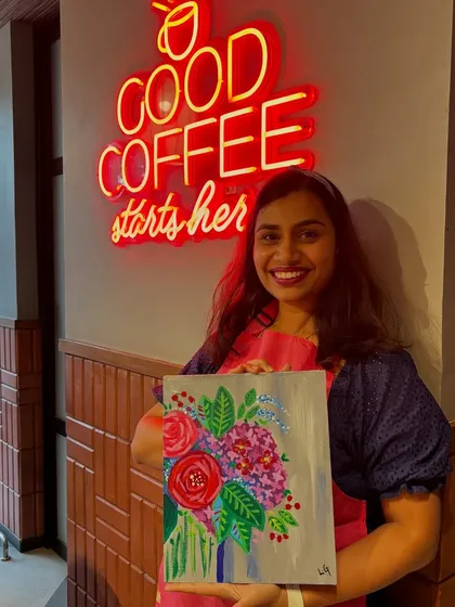 A participant posing with her finished floral painting in front of a neon sign at a cafe. The trendy locations add to the fun experience.