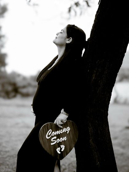 A serene black and white portrait of the mom-to-be leaning against a tree, holding a "Coming Soon" sign. The peaceful expression and natural setting create a timeless feel.