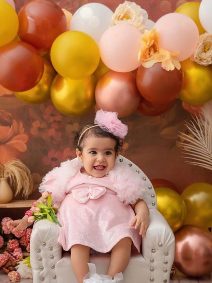 A cheeky little grin from the birthday girl, sitting in her chair surrounded by a beautiful arrangement of autumn-toned balloons.