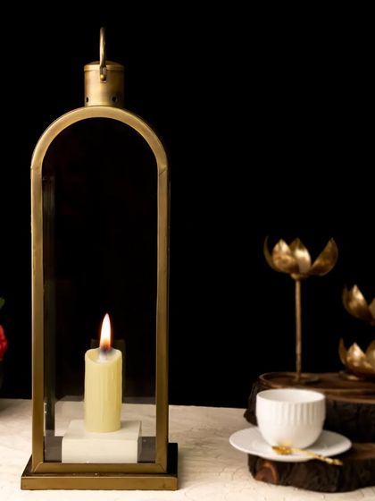 A tall, elegant brass lantern holds a single candle, creating a classic and timeless look. The dark background and additional brass decor add to the moody, sophisticated ambiance.