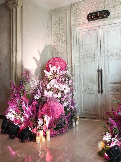 The entrance to a dreamy Mehendi celebration, where guests are greeted by lush arrangements of pink and purple flowers. A simple, elegant sign reads "All Because Two People Fell In Love," setting a romantic tone for the event.