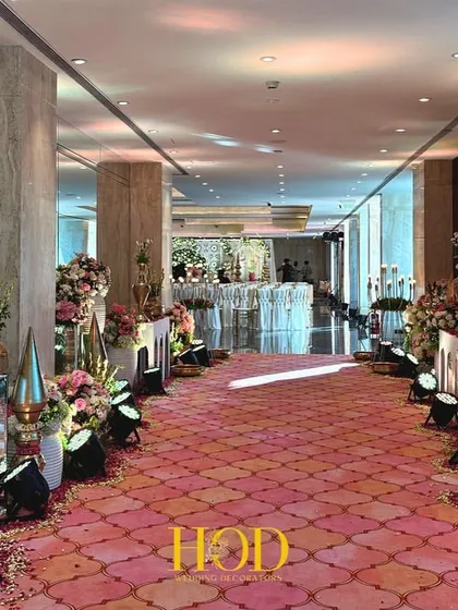 A long, dramatic walkway lined with traditional brass vessels, floral arrangements, and a patterned carpet, guiding guests through a journey of color and culture.