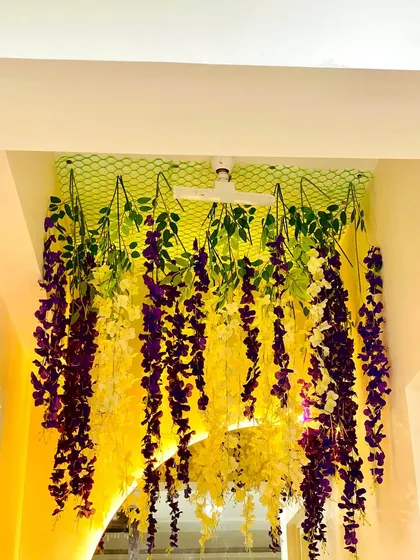 A close-up of a ceiling installation featuring hanging artificial wisteria flowers. The mix of purple, yellow, and white blooms creates a dramatic and elegant effect, perfect for event venues, boutiques, or even a statement piece in a home.