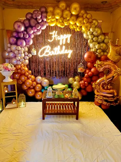We created this vibrant bedroom setup using a full ring of metallic rainbow-colored balloons. The shimmering curtain backdrop and central neon sign make for a festive and intimate birthday surprise.