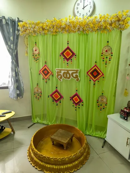 We can adapt our Haldi decor for any space, like this cozy living room setup. We used a light green curtain backdrop with traditional hangings, a wooden 'हळद' sign, and our signature golden Urli to create a festive corner.