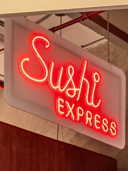 A close-up of the 'Sushi Express' neon sign. The retro-style script and bright red color give it a bold, playful character that perfectly suits the restaurant's theme.