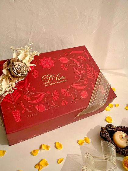 Our premium red Diwali hamper box, elegantly styled with scattered flower petals and a lit diya, capturing the festive spirit.
