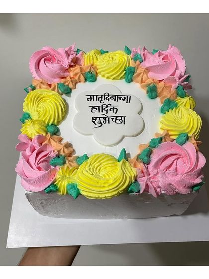 A beautiful square cake with a message in Marathi for Mother's Day. The floral piping in yellow and pink adds a bright, cheerful touch.
