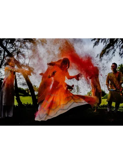 A bride is surrounded by a cloud of red and orange powder during a pre-wedding celebration. The dramatic lighting and motion create a powerful and artistic image of festive joy.