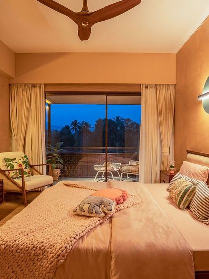 This bedroom uses a softer, lighter color palette while maintaining the home's earthy feel. The room opens onto a balcony, and the warm lighting from the modern wall sconce creates a serene ambiance in the evening.