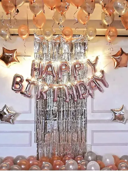 A chic birthday decoration with a silver fringe backdrop. Rose gold 'Happy Birthday' foil letters, matching ceiling balloons, and star accents create a trendy and festive look.