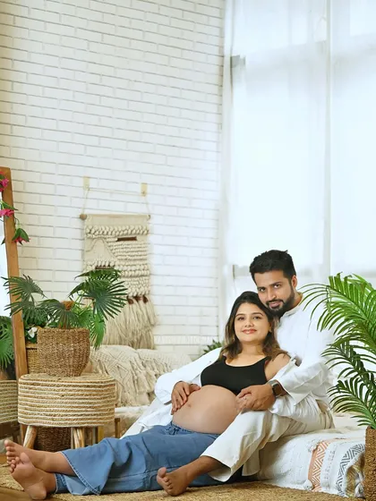 A full-length view of a couple during their boho-style maternity photoshoot, showcasing the comfortable and aesthetic studio setup with natural textures and plants.