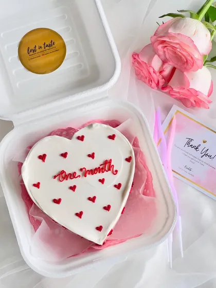 Simple, sweet, and perfect for anniversaries. This heart-shaped bento cake celebrates a one-month milestone with clean red piping and tiny hearts on a smooth white buttercream canvas, presented with fresh roses.