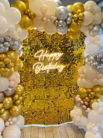 A glamorous birthday party with a gold sequin wall. The white, gold, and silver balloon arch and 'Happy Birthday' neon sign create a luxurious and celebratory atmosphere.
