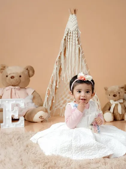 A sweet, seated portrait of the birthday girl in her white dress, surrounded by her furry teddy bear friends in a warm and inviting setting.