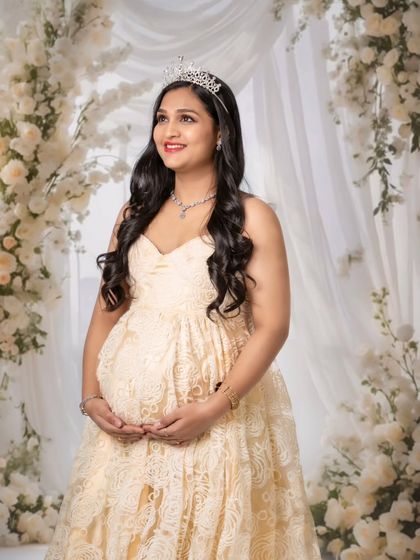 A close-up portrait of a radiant mom-to-be wearing a tiara, surrounded by a dreamy wall of white flowers.