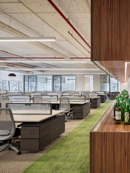 A view across the open workstations at the APT office, with a pantry and breakout area in the background. The layout is designed for easy flow and interaction, with green carpet paths visually guiding movement through the space.